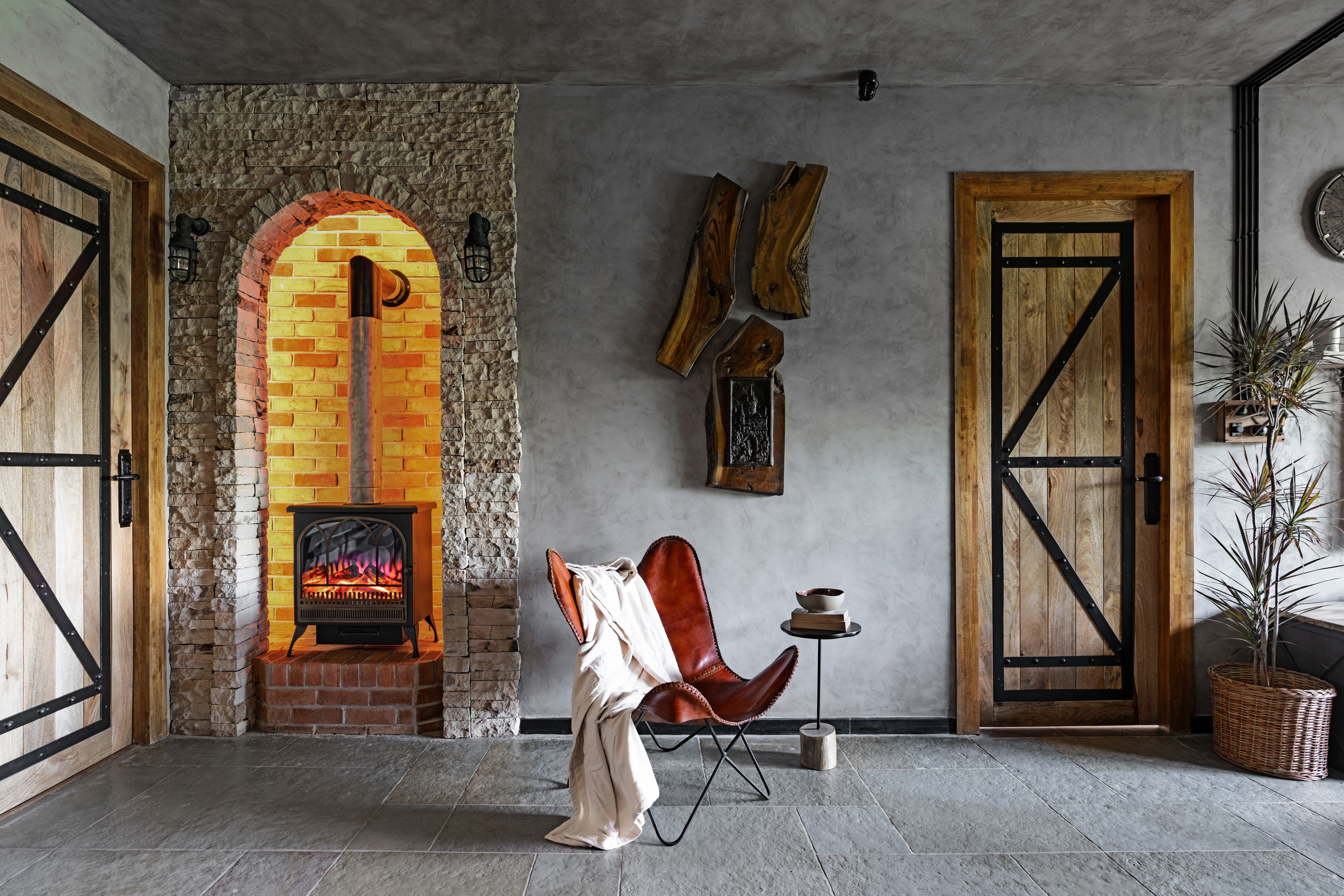 Hallway corner with a black stove in an arched stone niche, a tan leather butterfly chair draped with a cream throw, a small black side table with a bowl and books, live-edge timber pieces mounted on the plaster wall, and timber barn doors either side.